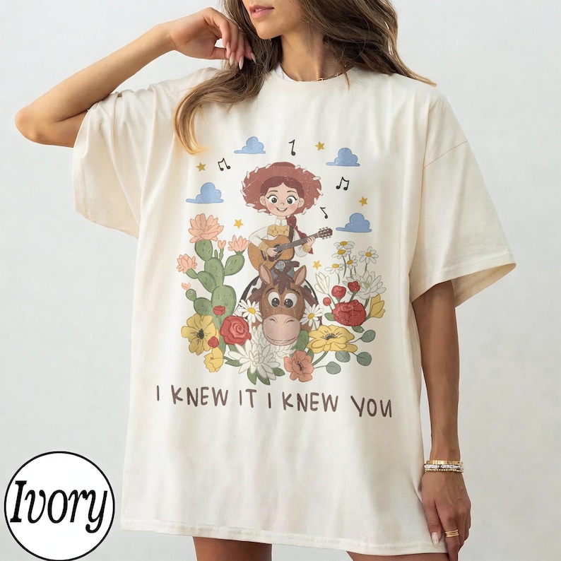 May include: An ivory t-shirt featuring a graphic print of a cartoon character with red hair playing a guitar while riding a brown horse. The design includes colorful flowers, cacti, blue clouds, musical notes, and the text I KNEW IT I KNEW YOU.