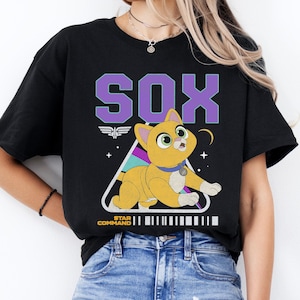 Op de afbeelding: Zwart T-shirt met een cartoon illustratie van Sox, een gele kat met groene ogen, uit de film Lightyear. Het woord "SOX" staat in paarse blokletters boven de kat. Op het shirt staan ook de woorden "STAR COMMAND".