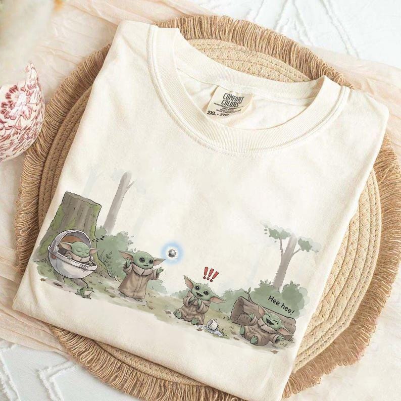 May include: Cream-colored t-shirt featuring a cartoon of a green alien character in various poses. The graphic includes the text "Hee hee!" and other visual elements. The shirt is folded on a woven mat.