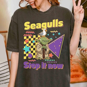May include: Dark grey t-shirt with a graphic design featuring a cartoon Yoda character with a seagull on his head. The text "Seagulls Stop it now" is in yellow and purple.