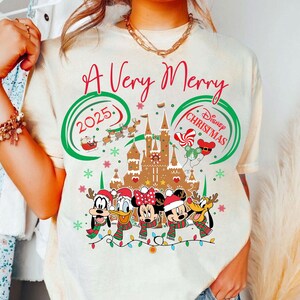 Può includere: T-shirt color crema con un design natalizio Disney festivo. La maglietta presenta la scritta "A Very Merry Christmas 2025" in rosso e verde, insieme a personaggi dei cartoni animati e un castello di pan di zenzero. I personaggi indossano cappelli e sciarpe di Babbo Natale.