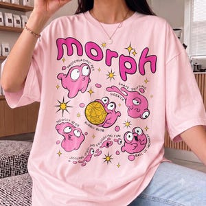 Op de afbeelding: Een lichtroze t-shirt met een grote afbeelding met het woord "morph" in dikke roze letters. Het ontwerp bevat verschillende roze, klodderachtige wezens met labels zoals "protoplasmic pet", "shape-shifting sidekick", "bubbly blob", "noogly glob", "jiggling", "squiggling fun" en "ball of machinery".