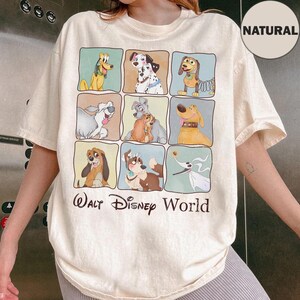 Op de afbeelding: Crèmekleurig T-shirt met een raster van cartoonhondenfiguren in kleurrijke vierkanten. Het shirt heeft de woorden "Walt Disney World" in een donkerbruin lettertype. Een cirkelvormig "NATURAL" label is zichtbaar.
