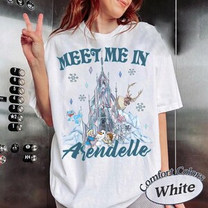 May include: White t-shirt with a graphic featuring a castle, reindeer, and characters from a popular animated movie. The text "Meet Me In Arendelle" is printed in blue script. The shirt is labeled "Comfort Colors White".