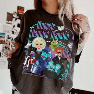 May include: Dark gray t-shirt featuring a colorful graphic of Muppets characters with the text "Muppets Haunted Mansion". The design includes Miss Piggy, Kermit, Gonzo, and other Muppets in a spooky setting.
