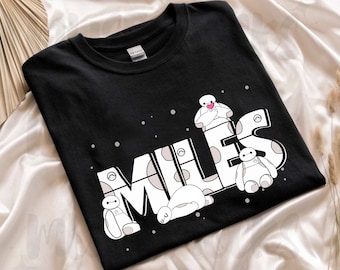 Op de afbeelding: Zwart T-shirt met de naam "MILES" in het wit, versierd met cartoon Baymax-personages. De letters zijn voorzien van stippen en het ontwerp bevat kleine witte stippen.
