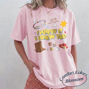 May include: A light pink short-sleeve t-shirt featuring a graphic print with the text I KNEW IT I KNEW YOU, infinity and beyond, brown cowboy boots, a yellow sheriff star, alphabet blocks, a red hat, and a colorful beaded necklace design.