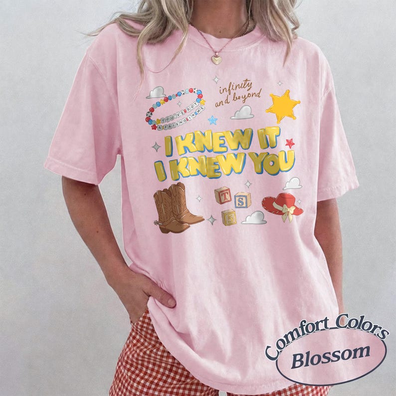 May include: A light pink short-sleeve t-shirt featuring a graphic print with the text I KNEW IT I KNEW YOU, infinity and beyond, brown cowboy boots, a yellow sheriff star, alphabet blocks, a red hat, and a colorful beaded necklace design.