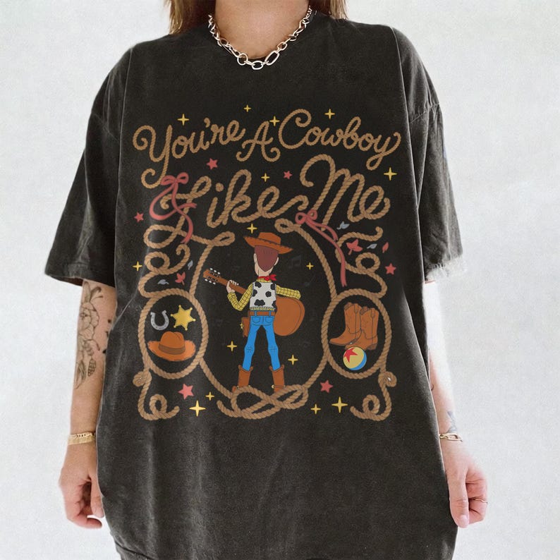 May include: A charcoal gray short-sleeve t-shirt featuring a graphic print of a cowboy character playing a guitar. The design includes a rope border, stars, a horseshoe, a hat, and boots, with the text "You're A Cowboy Like Me" in script.