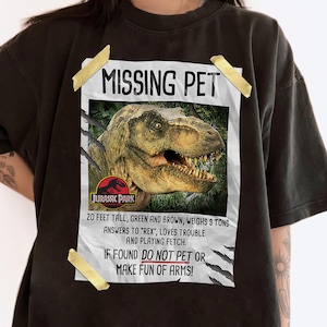 May include: Black t-shirt featuring a "Missing Pet" poster design with a dinosaur graphic and the Jurassic Park logo. The poster text includes details about the missing dinosaur, including its size, weight, and behavior.