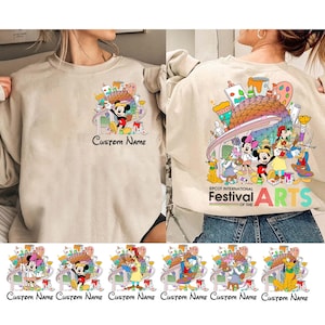 May include: A beige sweatshirt with a colorful graphic print of Mickey Mouse and other Disney characters at Epcot's International Festival of the Arts. The text "Custom Name" appears below the graphic. The back of the sweatshirt features a larger version of the graphic with the text "EPCOT International Festival of the Arts".