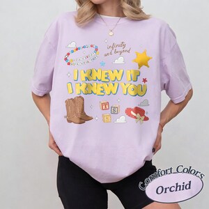 May include: An orchid-colored t-shirt featuring a graphic print with the text I KNEW IT I KNEW YOU, infinity and beyond, and You've got a friend in me. The design includes illustrations of brown cowboy boots, alphabet blocks, a hat, and a star.