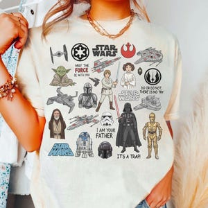 May include: Cream-coloured t-shirt featuring various Star Wars characters and quotes. The design includes Yoda, Darth Vader, Luke Skywalker, and the text "I am your father." Other phrases include "May the Force be with you" and "It's a trap!"