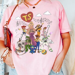 May include: A light pink t-shirt featuring a graphic print of various animated characters, a vintage camera, musical notes, and a red heart containing the letters TS. The design includes figures in colourful outfits, a ball, and white clouds.