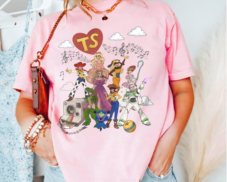 May include: A light pink t-shirt featuring a graphic print of various animated characters, a vintage camera, musical notes, and a red heart containing the letters TS. The design includes figures in colourful outfits, a ball, and white clouds.