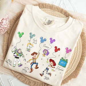 Op de afbeelding: Cr&egrave;mekleurige sweatshirt met stripfiguren Buzz Lightyear, Woody en Jessie, elk met een Mickey Mouse-ballon. Het shirt bevat de namen van de personages en andere tekst. Het shirt ligt op een geweven mat.