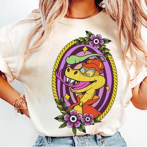 Pode incluir: Uma t-shirt de cor creme com um gráfico de uma cobra amarela a usar um chapéu e uma capa vermelhos. A cobra está emoldurada por uma borda de corda amarela, círculos concêntricos roxos e flores roxas com folhas verdes. O design é vibrante.