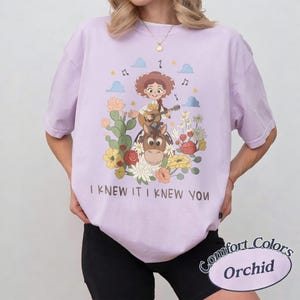 May include: A light purple t-shirt featuring a graphic of a cowgirl playing a guitar while riding a horse, surrounded by cacti and flowers. The text "I KNEW IT I KNEW YOU" is printed below the illustration. The shirt has short sleeves and a crew neck.