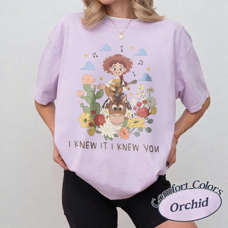 May include: A light purple t-shirt featuring a graphic of a cowgirl playing a guitar while riding a horse, surrounded by cacti and flowers. The text "I KNEW IT I KNEW YOU" is printed below the illustration. The shirt has short sleeves and a crew neck.