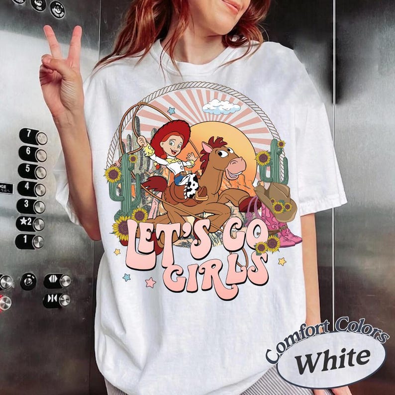 May include: White t-shirt with a cartoon graphic of a cowgirl riding a horse, surrounded by sunflowers and cacti. The text "LET'S GO GIRLS" is displayed in pink. The shirt is labeled "Comfort Colors White".