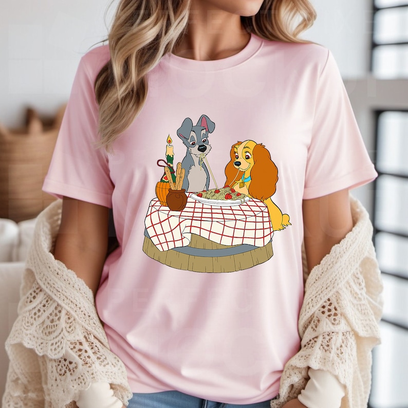 May include: A pink t-shirt with a cartoon image of Lady and the Tramp sharing a plate of spaghetti. The image is in a vintage style with a red and white checkered tablecloth.