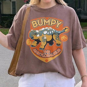 Könnte beinhalten: Ein braunes T-Shirt mit kurzen Ärmeln und einer Grafik eines Ankylosaurus-Dinosauriers im Cartoon-Stil. Der Text lautet BUMPY THE ANKYLOSAURUS in Orange und Gelb. Das Design enthält ein kleines Dinosaurier-Ei und ein kreisförmiges Symbol.