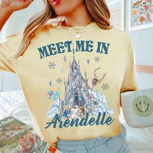 May include: Yellow t-shirt with the text "MEET ME IN Arendelle" in blue. The graphic features a castle, characters, and snowflakes. The shirt is a light yellow color.
