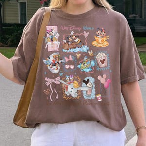 May include: Brown t-shirt featuring a collage of Disney characters and icons, including Mickey and Minnie Mouse, Dumbo, and popcorn. The shirt has the text "Walt Disney World" and "The Most Magical Place on Earth."