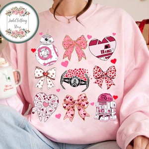 Puede incluir: Sudadera rosa con un tema de San Valentín con personajes de Star Wars como R2-D2, BB-8 y Darth Vader. Los personajes están rodeados de corazones y lazos.