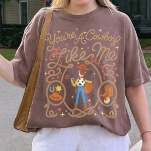 May include: A brown short-sleeve t-shirt featuring a graphic of a cowboy character playing a guitar. The text "You're A Cowboy Like Me" is printed above the character, surrounded by lasso ropes, stars, a horseshoe, a hat, boots, and a ball.