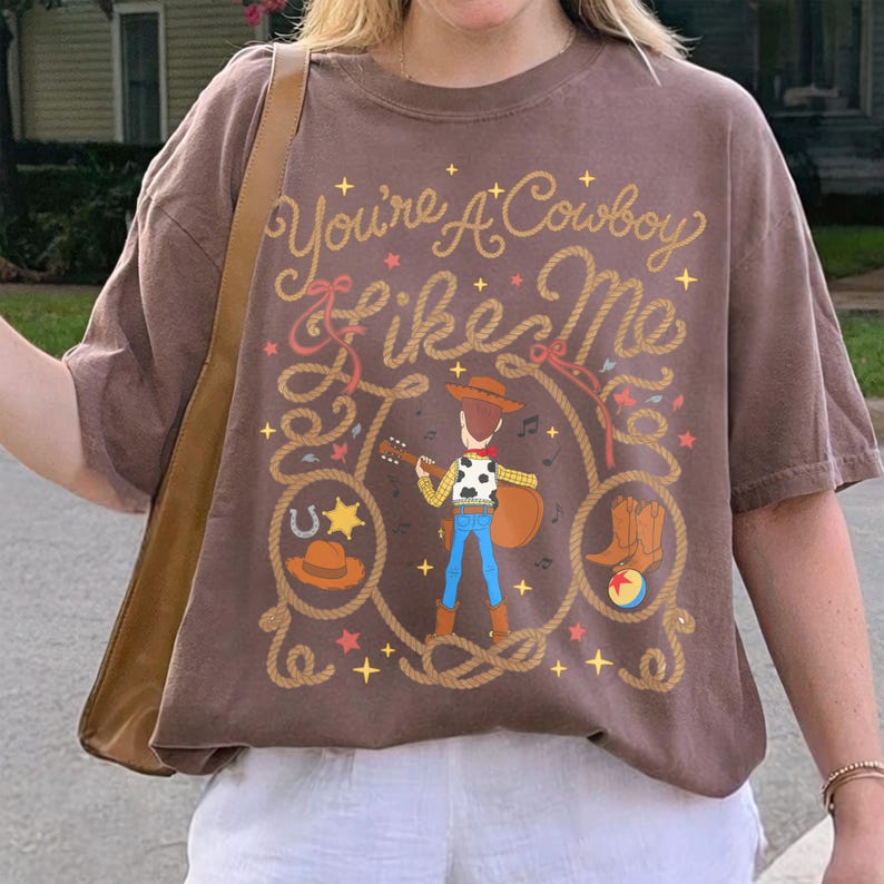 May include: A brown short-sleeve t-shirt featuring a graphic of a cowboy character playing a guitar. The text "You're A Cowboy Like Me" is printed above the character, surrounded by lasso ropes, stars, a horseshoe, a hat, boots, and a ball.
