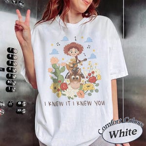 May include: A white t-shirt featuring a graphic of a cartoon cowgirl playing a guitar while riding a horse, surrounded by cacti, flowers, clouds, and musical notes. The text "I KNEW IT I KNEW YOU" is printed in brown below the illustration.