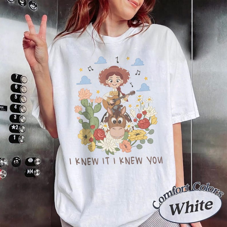 May include: A white t-shirt featuring a graphic of a cartoon cowgirl playing a guitar while riding a horse, surrounded by cacti, flowers, clouds, and musical notes. The text "I KNEW IT I KNEW YOU" is printed in brown below the illustration.