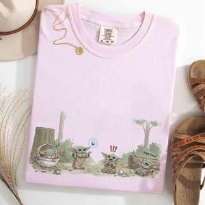 May include: A light pink Comfort Colors t-shirt featuring a cartoon illustration of Grogu from The Mandalorian. The design shows Grogu in various poses with the text "Hee hee!" and a blue orb. A gold necklace and brown sandals are also visible.