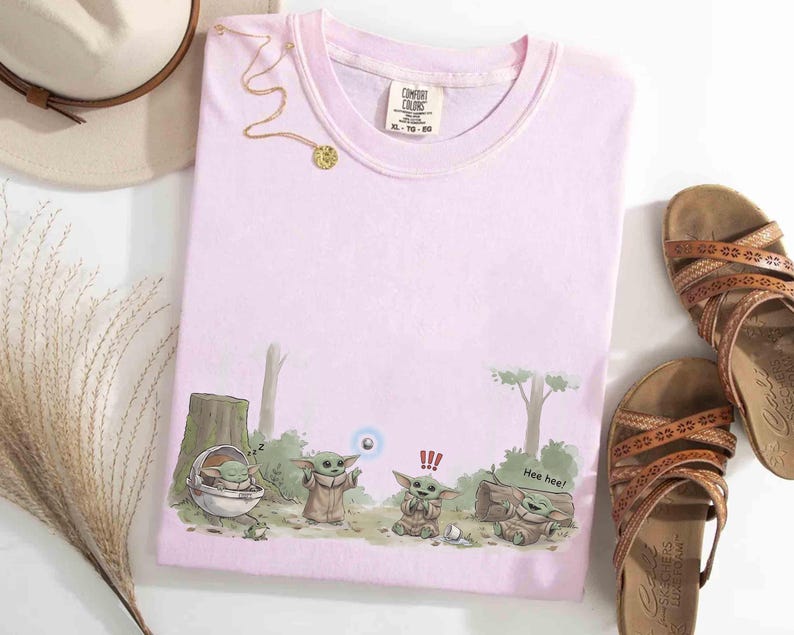 May include: A light pink Comfort Colors t-shirt featuring a cartoon illustration of Grogu from The Mandalorian. The design shows Grogu in various poses with the text "Hee hee!" and a blue orb. A gold necklace and brown sandals are also visible.