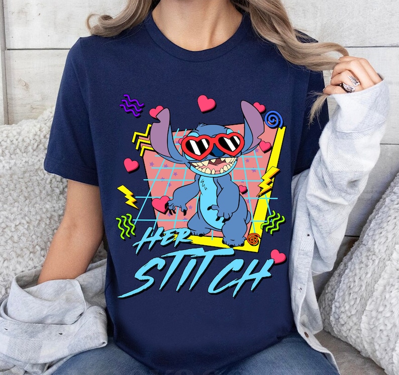 Peut inclure: Un t-shirt bleu marine avec un motif r&eacute;tro de Stitch de Lilo & Stitch portant des lunettes de soleil en forme de c&oelig;ur. Le motif comprend des accents roses, jaunes et bleus. Le texte "Her Stitch" est imprim&eacute; en gras et en style stylis&eacute; sous le motif.
