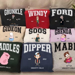 Op de afbeelding: Een collectie van negen gevouwen sweatshirts in diverse kleuren, elk voorzien van een Gravity Falls-personage, de naam in grote letters in varsity-stijl, de tekst EST 2012 en de woorden GRAVITY FALLS gedrukt in witte of zwarte inkt.