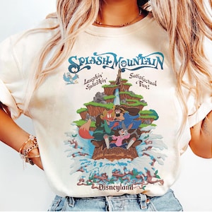 Puede incluir: Una camiseta de color crema con un gráfico de Splash Mountain que muestra a Br'er Rabbit, Br'er Fox y Br'er Bear en un tronco. El texto dice Splash Mountain, Laughin' Splashin' Satisfactual Fun! y Disneyland en azul oscuro y marrón.