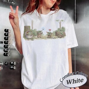 May include: White Comfort Colors t-shirt featuring a cartoon illustration of Grogu in various poses. The design includes Grogu in a pod, standing, and sitting near a log with the text "Hee hee!".