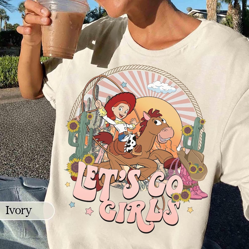 May include: Ivory-colored t-shirt featuring a cartoon cowgirl riding a horse with a lasso, set against a desert landscape with sunflowers and cacti. The text "LET'S GO GIRLS" is in pink script. A cowboy hat and boots are also shown.