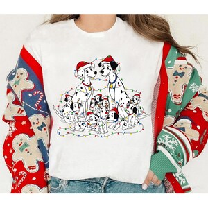 May include: White t-shirt featuring a graphic of Dalmatian dogs wearing Santa hats, surrounded by Christmas lights. The shirt is worn with a red, white, and green patterned sweater. The sweater has gingerbread men and snowflake designs.