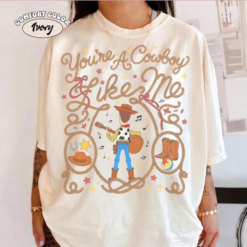 May include: An ivory t-shirt featuring a graphic of a cowboy character playing a guitar, surrounded by a rope lasso design. The text reads "You're A Cowboy Like Me" in brown cursive script, accompanied by small stars, musical notes, a hat, and boots.