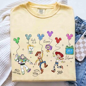 Op de afbeelding: Een lichtgeel t-shirt met stripfiguren Buzz Lightyear, Woody en Jessie, elk met een Mickey Mouse-vormige ballon. Het shirt bevat de tekst "Buzz Lightyear", "Woody" en "Jessie". Andere personages zijn "Smarty Pants", "Snappy" en "Lilypad".