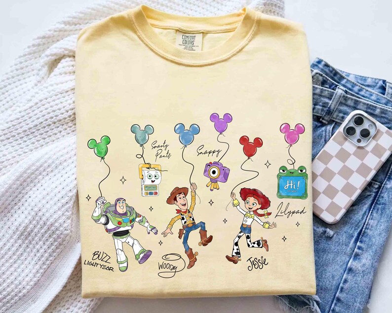 Op de afbeelding: Een lichtgeel t-shirt met stripfiguren Buzz Lightyear, Woody en Jessie, elk met een Mickey Mouse-vormige ballon. Het shirt bevat de tekst "Buzz Lightyear", "Woody" en "Jessie". Andere personages zijn "Smarty Pants", "Snappy" en "Lilypad".