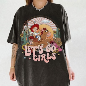 May include: Dark gray t-shirt featuring a cartoon cowgirl riding a horse, with a sunset backdrop and cacti. The text "LET'S GO GIRLS" is displayed in pink and white. A cowboy hat and boots are also shown.