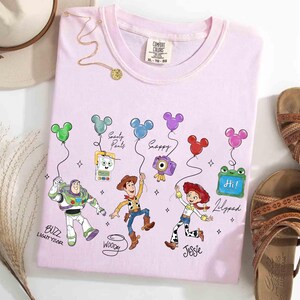 Op de afbeelding: Een lichtroze t-shirt met tekenfilmfiguren Buzz Lightyear, Woody, Jessie en anderen die Mickey Mouse-vormige ballonnen vasthouden. Het shirt heeft de tekst "BUZZ LIGHTYEAR", "Woody", "Jessie", "Lilypad", "Snappy" en "Soarty Pants". Bruine sandalen en een hoed liggen ernaast.