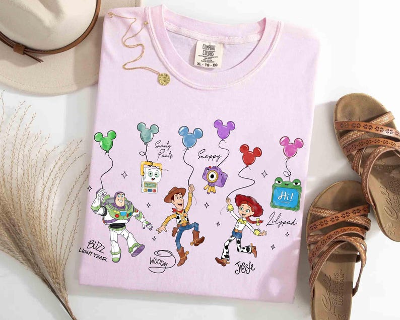 Op de afbeelding: Een lichtroze t-shirt met tekenfilmfiguren Buzz Lightyear, Woody, Jessie en anderen die Mickey Mouse-vormige ballonnen vasthouden. Het shirt heeft de tekst "BUZZ LIGHTYEAR", "Woody", "Jessie", "Lilypad", "Snappy" en "Soarty Pants". Bruine sandalen en een hoed liggen ernaast.