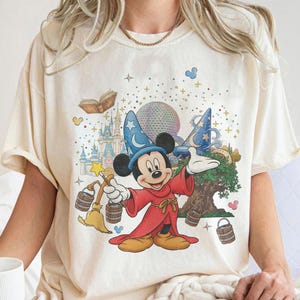 Peut inclure: Un t-shirt couleur crème avec une illustration de Mickey Mouse en sorcier portant une robe rouge et un chapeau bleu. Le motif comprend un château, une sphère géodésique, un arbre, des seaux flottants, un balai, un livre ouvert et des étoiles.