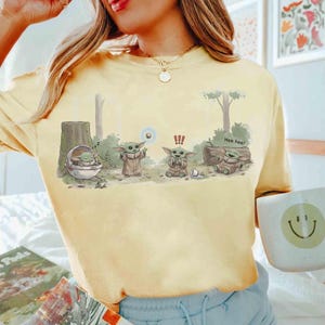 May include: Yellow t-shirt featuring a cartoon illustration of a character from a popular science fiction franchise. The graphic depicts the character in various poses with the text "Hee hee!" and other symbols.