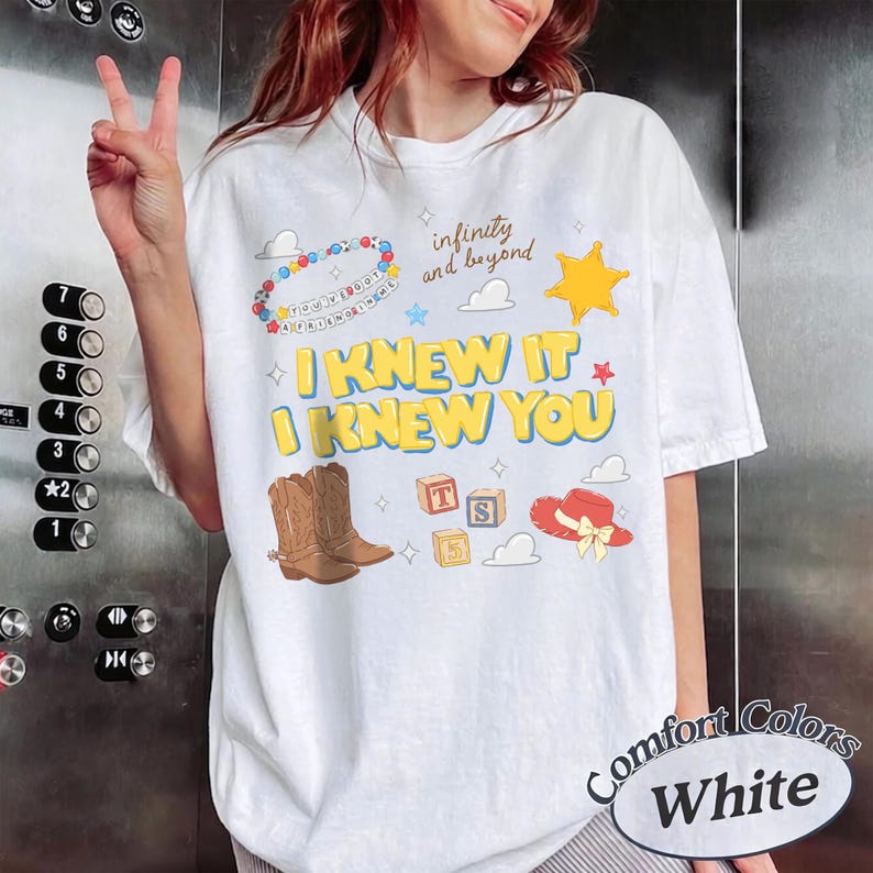 May include: A white t-shirt featuring a graphic print with the text "I KNEW IT I KNEW YOU" in yellow, "infinity and beyond" in brown, and "YOU'VE GOT A FRIEND IN ME" on a beaded necklace. Includes illustrations of brown cowboy boots, blocks, and a hat.